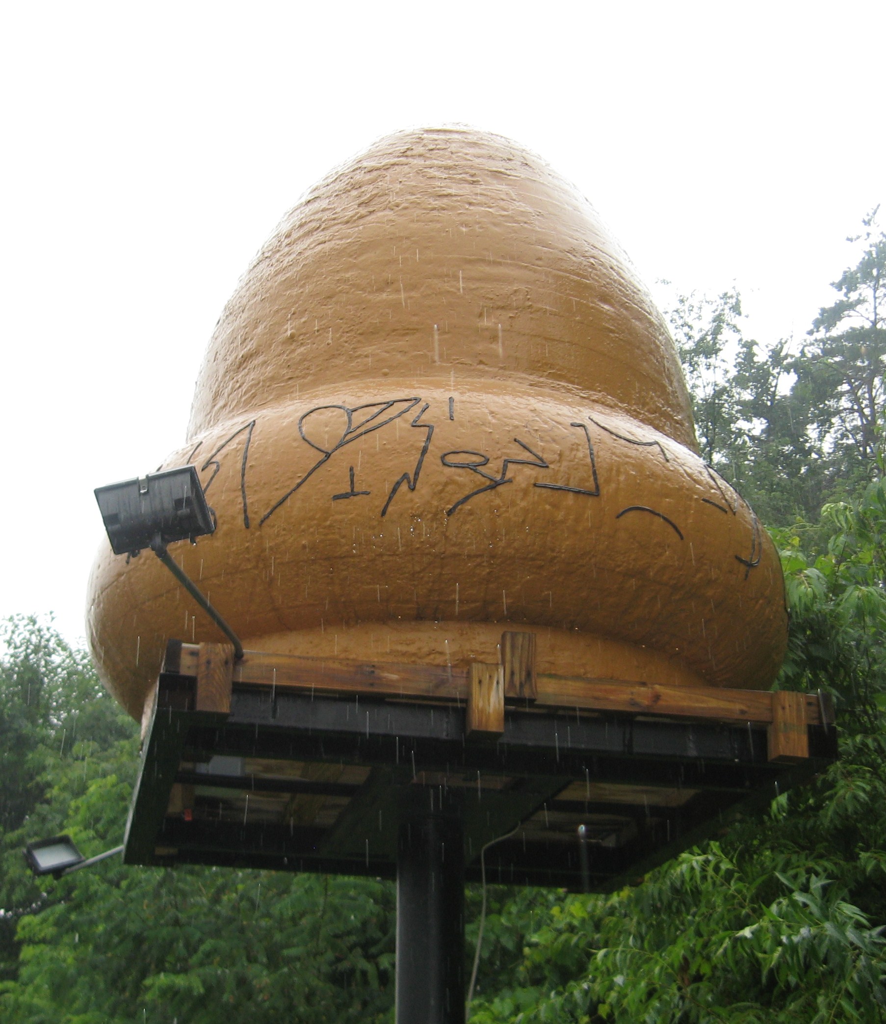 Photograph of the large bronze-colored acorn-shaped Kecksburg UFO replica mounted outdoors among green trees near the volunteer fire station.