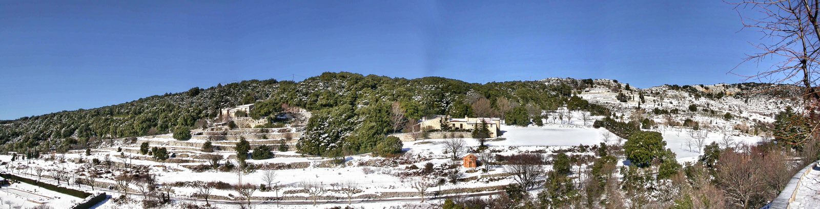 Wide winter panorama of dry-stone terraced hillsides under snow, with dark evergreen oaks, a few stone farm buildings and a bright blue sky.