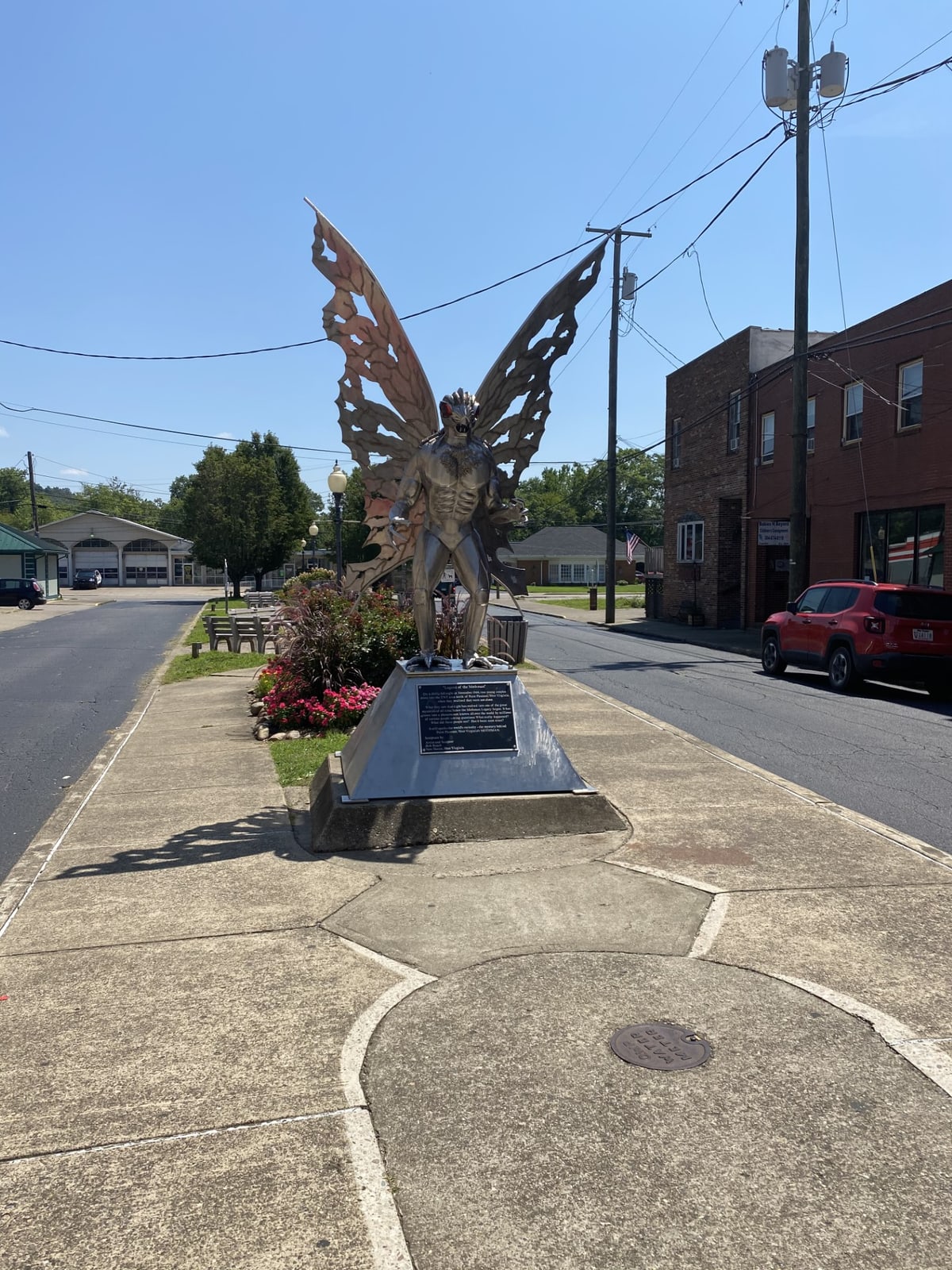 A polished stainless-steel statue of a winged, red-eyed humanoid on a plinth beside a small-town main street under a clear sky.
