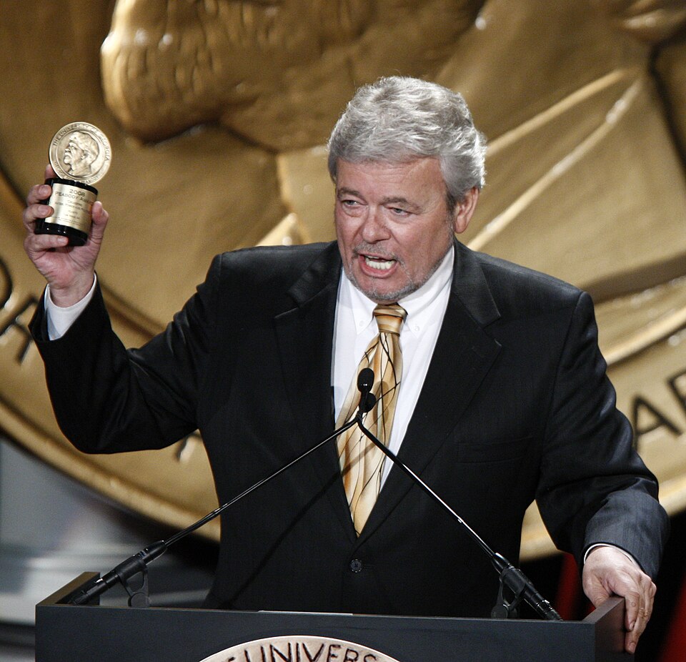 George Knapp in a suit holding a Peabody Award at a lectern.