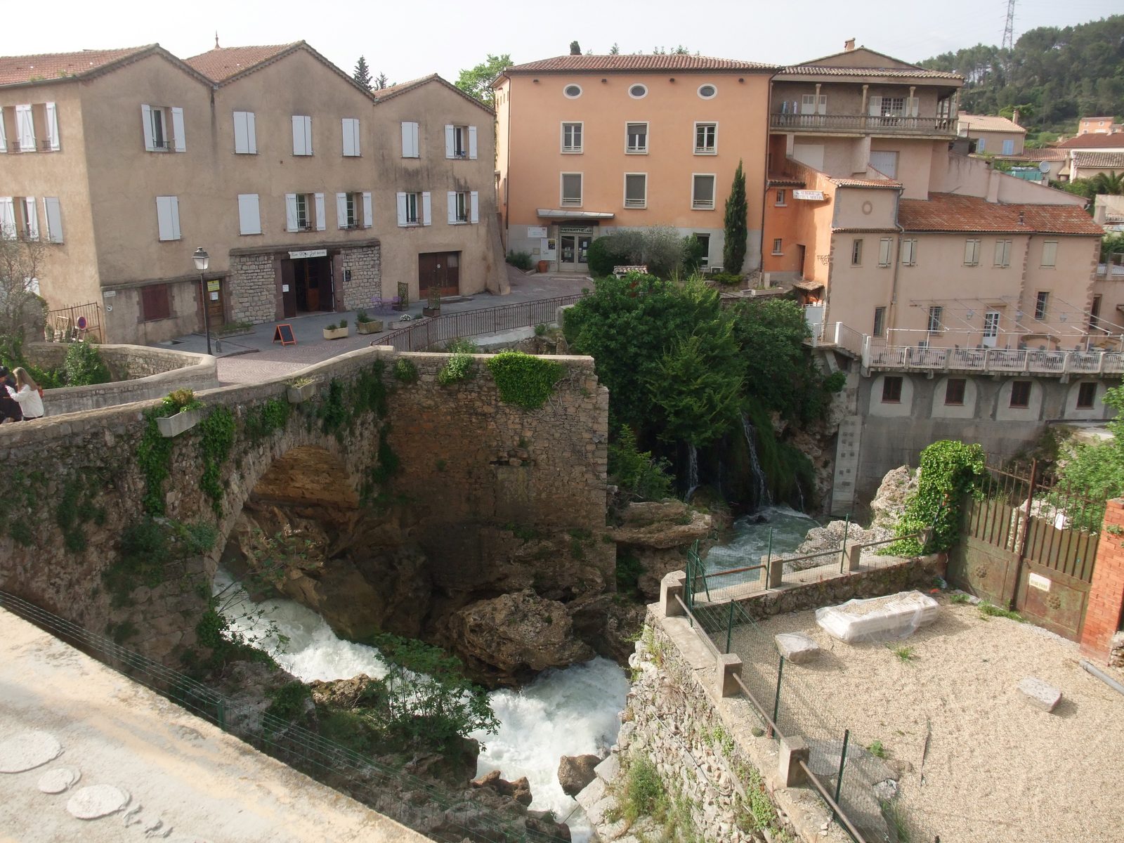 Photograph of the center of Trans-en-Provence: a white Nartuby cascade tumbling under an old stone bridge between ochre and cream houses with green shutters and terraces.