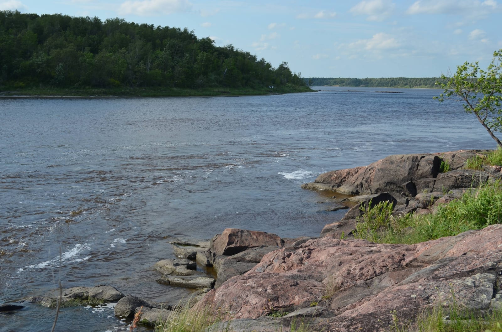Pink Precambrian granite shelving into the Whiteshell River under a summer sky, with boreal forest on the far bank.