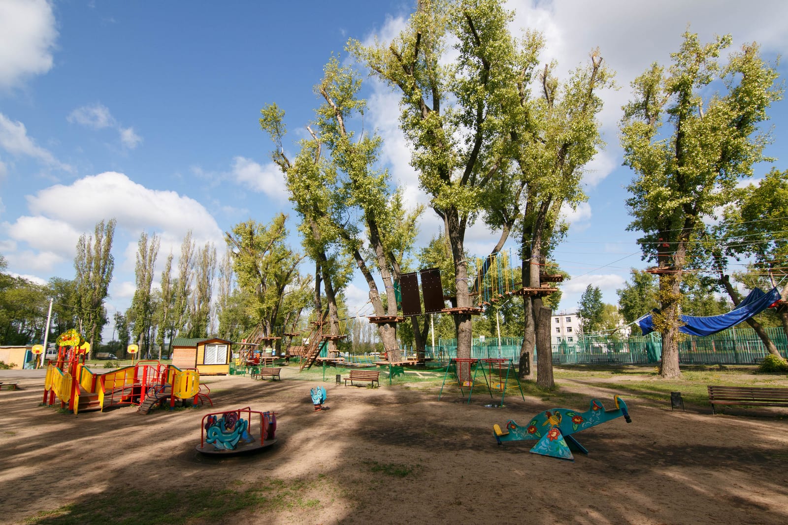 Um parquinho sob choupos altos no Parque Yuzhny, em Voronezh, com um percurso de cordas armado entre os troncos e blocos de apartamentos da era soviética atrás de uma cerca verde.